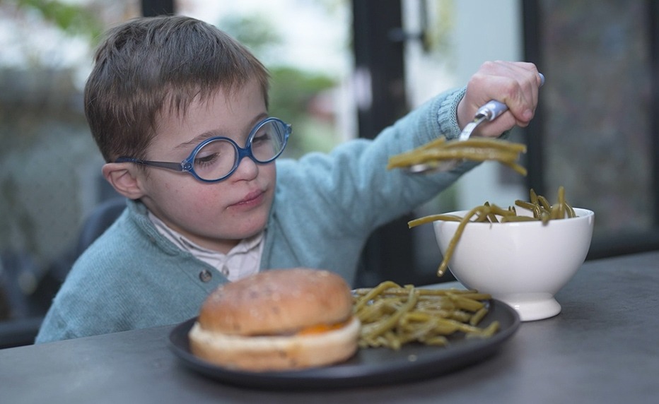 Petit garçon trisomique mangeant des haricots verts avec un hamburger